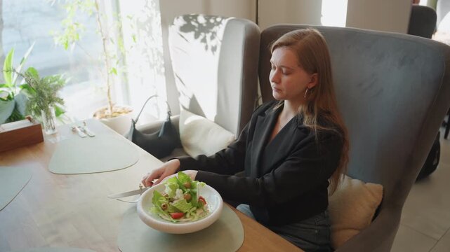 White businesswoman pausing with salad at cafe, executive on work break wearing blazer, handbag and phone on table, leafy salad in ceramic bowl, sunlit window, elegant chair, calm reflective mood,