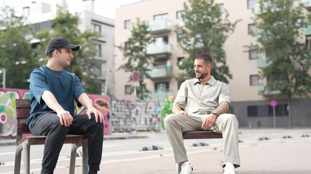 Two men converse on a city bench. One listens while the other gestures, hands held. Urban backdrop features graffiti and trees. Natural light casts soft shadows around them. Ideal for social, urban