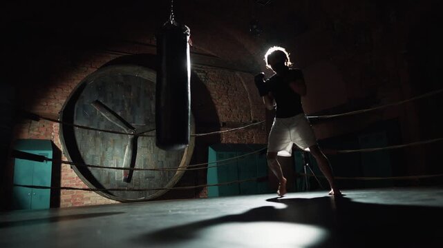 Cinematic shot in dark boxing club, male fighter practicing punches in ring. Professional sportsman training alone before important fight, future champion, silhouette of athletic man against soffit