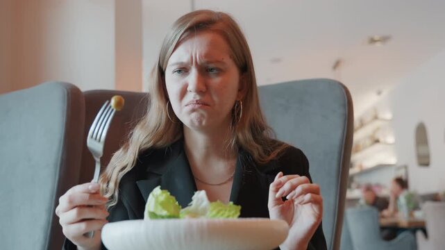 Caucasian woman disgusted tasting olive reaction in chic bistro, expressive grimace after unpleasant bite, customer inspecting salad quality, subtle tension in stylish blazer, interior lights