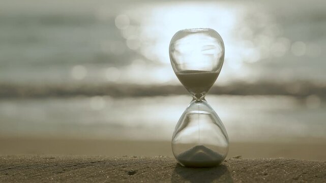 Glass hourglass on a sandy shore glowing in golden sunlight, representing time, waiting, deadlines, change, reflection, and the precious balance between past, present, and future.