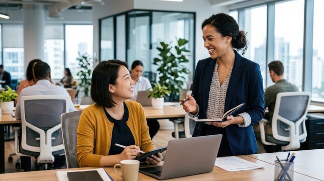 Two women are engaged in a collaborative discussion at a modern office desk. A senior businesswoman is guiding a younger colleague, who is working on a laptop.