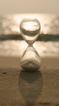 Hourglass standing on sandy beach during golden sunset with ocean waves in background. Concept of time passing, mindfulness, fleeting moments, life balance and peaceful coastal atmosphere in cinematic