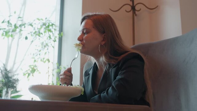 Caucasian businesswoman enjoying mindful lunch break windowlit cafe scene with caucasian woman in blazer holding forkful of lettuce like flower, bowl on table, plants in background, serene savoring,