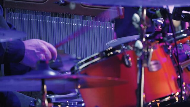 During a live jazz performance, a drummer beats out a rhythm on a drum kit. Bright purple and red stage lights create an energetic, atmospheric setting. Close-up with shallow depth of field