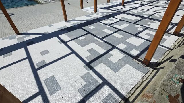 Modern outdoor flooring displaying a gray and white mosaic pattern, contrasting with strong, angular shadows cast by wooden support beams on a sunny day, creating an abstract design