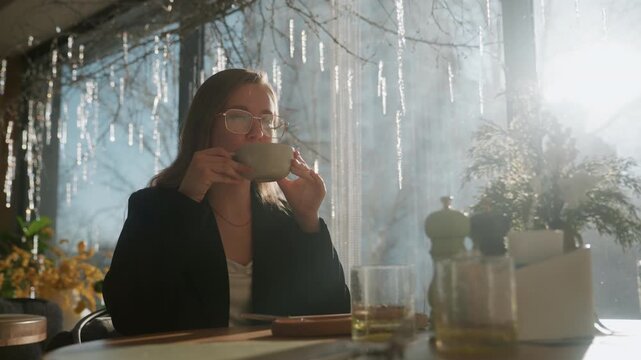 Sunlit cafe portrait white woman writer sipping cup, glasses catching lens flare, bright window backlight with plants and table setting, notebook and glass on table, serene reflective mood, slow