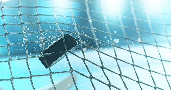Action sports photo of a hockey puck flying into the goal net during a scoring shot, with ice particles in the air and a hockey stick visible in the background, capturing speed and intensity.