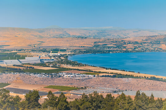 Horizontal wide angle eye level shot of the Sea of Galilee shoreline in Israel with agricultural fields, coastal buildings, and arid hills under a clear sunny sky