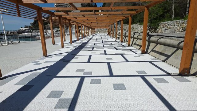 Long wooden pergola structure extending along a promenade, creating intricate shadow patterns on the patterned concrete path, leading the eye towards the horizon under a bright sunny sky