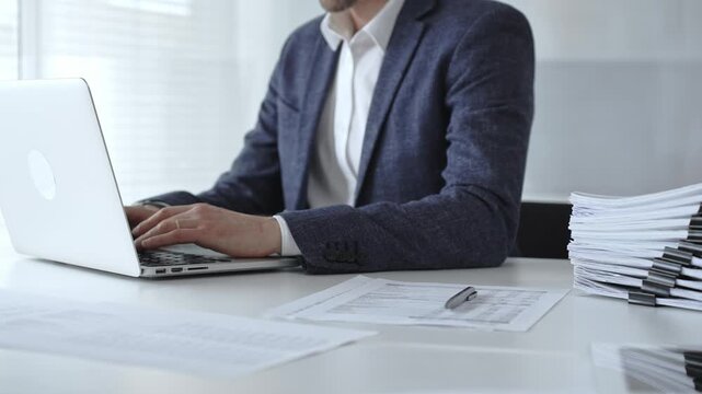 Businessman typing on laptop amid stacked documents and reports, focused on financial data and paperwork in a modern office, conveying productivity, management, and analysis. Audit and taxes