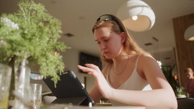 White woman focused typing on tablet, intense expression with pendant necklace, lamp and glass vase in foreground, busy cafe interior with warm evening light, multitasking on email and spreadsheets