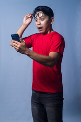 Fototapeta premium An expression of shock or disbelief is seen as an Asian man from Indonesia holds his cell phone and raises his glasses to his forehead. Isolated against a blue background.