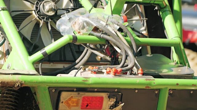 A close-up of a custom off-road buggy. A bright green roll cage encloses the winch, wiring, and cooling fans. A broken taillight and dirt indicate intense competition and extreme racing