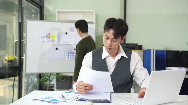 Business documents on office table with tablet and laptop computer and chart and two colleagues discussing data in office.