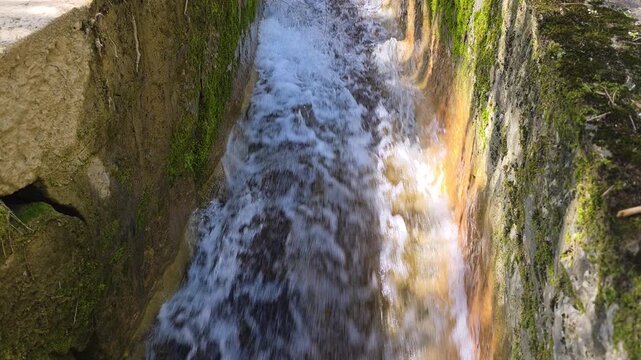 Rapid water rushing through a narrow human-made channel flanked by textured stone walls cloaked in vibrant green moss, sunlight highlighting motion, texture, and fresh natural energy