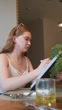 Young woman designing floral table layout with pen and clipboard, greenery and glassware on table creating botanic mood indoor cafe lighting, thoughtful expression suggesting event planning, stylist