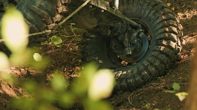 A close-up of a large, dirty tire and the suspension of a 4x4 SUV. The vehicle navigates rough forest terrain during an extreme off-road competition. The winch cable rubs against the studded tire