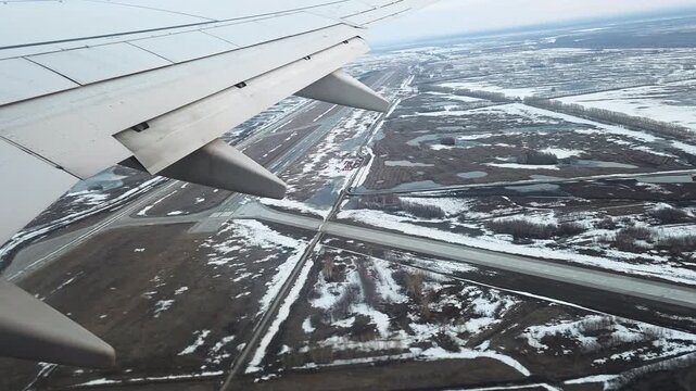 Airplane wing with flaps extended approaching a snowy, patchy runway and puddled fields below in a cold, overcast winter aerial view during landing approach