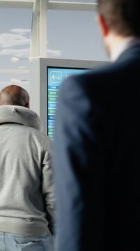 Vertical video Diverse tourists with trolleys in airport checking flight status information on screen. Passengers walking in airplane terminal, looking at flight timetable on electronic announcement