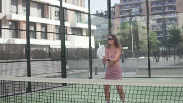 Active young woman executing a powerful smash shot in a padel tennis match. Female player in sunglasses demonstrating strength, precision, and dynamic movement on a modern outdoor court. Concept of a
