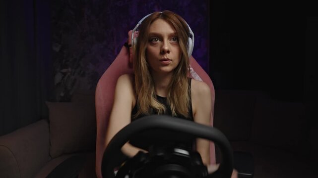 Woman driver concentrating intensely on steering wheel, headset fitted, leaning forward in pink gaming chair with focused stare