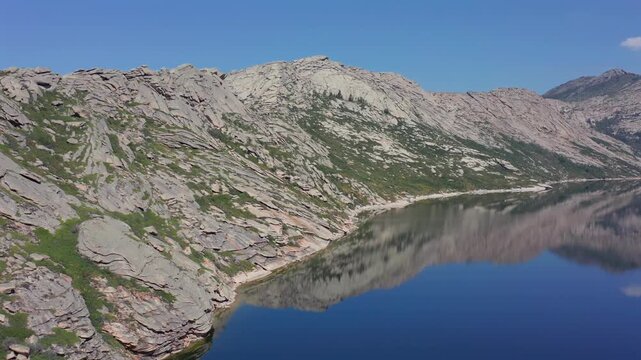 Aerial glide along rocky shoreline of Kolzhinkor lake in Kazakhstan, drone moves parallel to calm water with clear reflections, textured granite hills and untouched natural scenery.