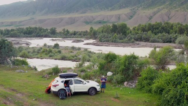 Car parked near riverbank as drone lifts upward revealing Chilik River valley landscape with flowing water, green vegetation and mountains in Kazakhstan travel destination