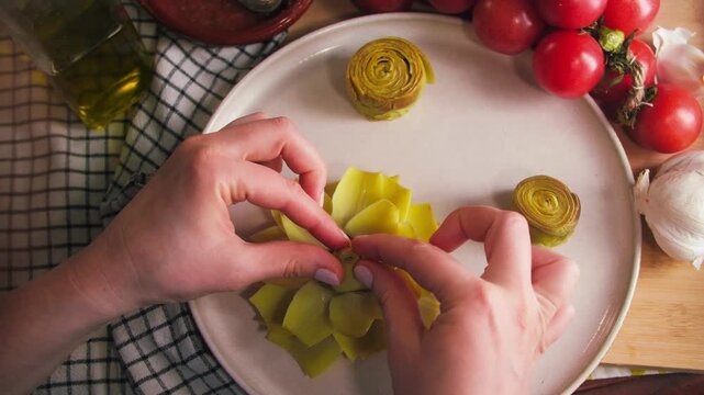 Close-up of hands opening artichoke petals and flattening the flower to prepare it for frying on both sides. The scene shows the cooking process of traditional Spanish dish las flores de alcachofa