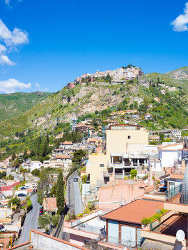 Scenic view of Castelmola village perched on rocky cliff above Taormina. Mediterranean architecture, red tile roofs, winding roads and lush green hills under clear blue sky in Sicily, Italy.