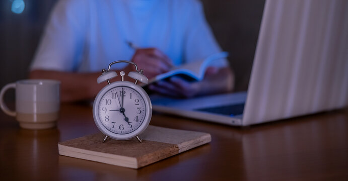 Person staying focused on remote work late at night or early morning, writing in a notebook near an alarm clock and laptop, highlighting dedication and productivity