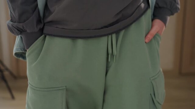 Green cargo pants pocket closeup hands tucked in pockets, textured knit fabric and functional drawstring on display, casual utility styling in hallway, soft window light highlights seams and pocket