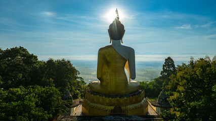 Obraz premium A majestic golden Buddha statue surrounded by lush green trees, set against a stunning backdrop of rolling mountains and a clear blue sky. This serene landscape captures the harmony of spirituality