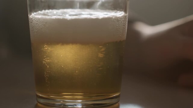 Closeup settled beer glass with foam, delicate bubbles rising in warm backlight, blurred arm resting in background, cozy minimal tabletop scene with serene moody atmosphere ideal for beverage