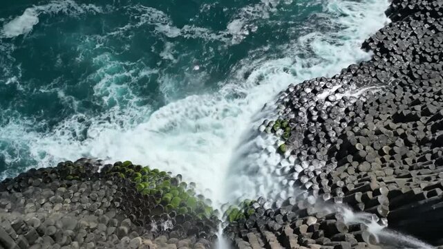 Aerial view of ocean waves crashing against a rock formation of hexagonal basalt columns with green moss