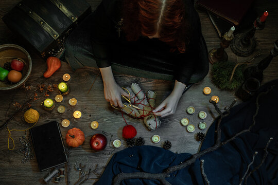 Witch wizard woman witchcrafting, performing ritual with handmade voodoo doll, sitting on the floor among candles, pumpkins. Magician Halloween night