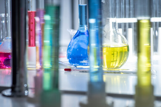 Chemical laboratory shelves with glass flasks and beakers under blue light, blurred foreground glassware, modern science workspace background with copy space.