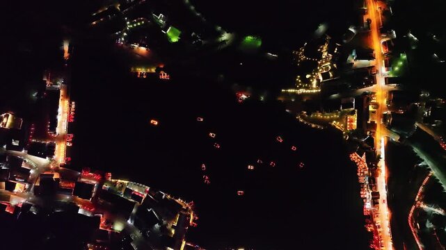A stunning aerial view of a lakeside village at night, illuminated with warm golden lights. Floating lanterns and boats create a mesmerizing pattern on the dark water, Baan Rak Thai Chinese style