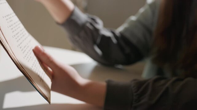Hands holding aged book pages, sunlit student turning fragile manuscript on white table, warm morning light, soft shadows, careful fingers tracing text, quiet archival study mood