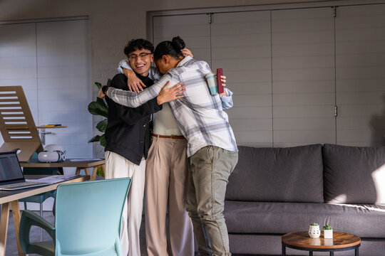 Three friends hugging, sharing warm moment in living room with laptop, travel mug, succulents