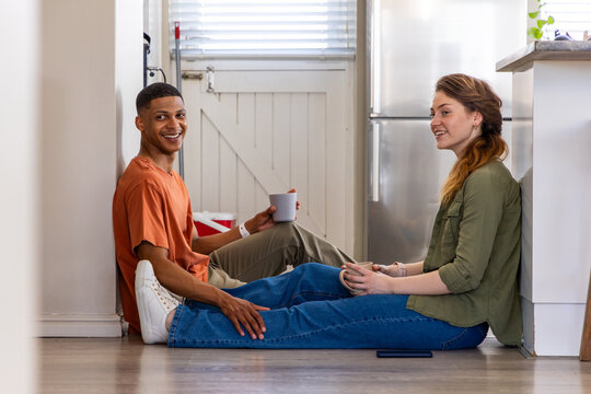 Couple sitting on kitchen floor facing each other, holding ceramic mugs near smartphone