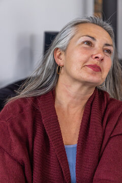 Senior woman sitting in living room wearing maroon cardigan and gold hoop earrings, looking upward
