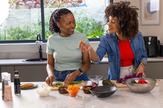Two African American women chopping and tasting vegetables at marble kitchen island with bowls