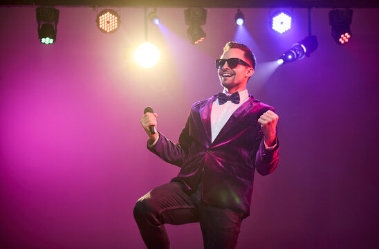 Singer presenter showman host performer with microphone. A confident man on stage under concert spotlight engages the crowd with upbeat energy. Concept of celebration, show, and entertainment.