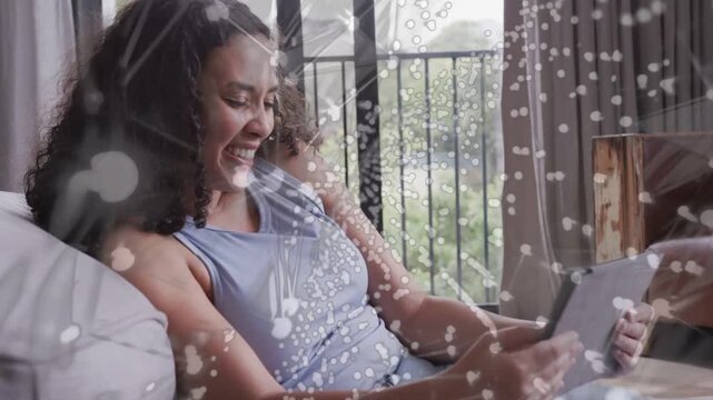 Woman tapping tablet on couch by balcony, leaning back laughing, dots overlaying tech relaxing mood