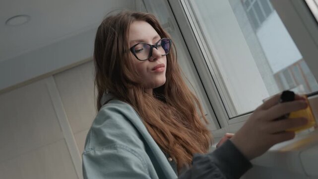 Caucasian woman drinking juice presenting product sample by window, glasses reflecting daylight, cheerful influencer demo with bottle sip and handheld device reveal, candid smile and approachable