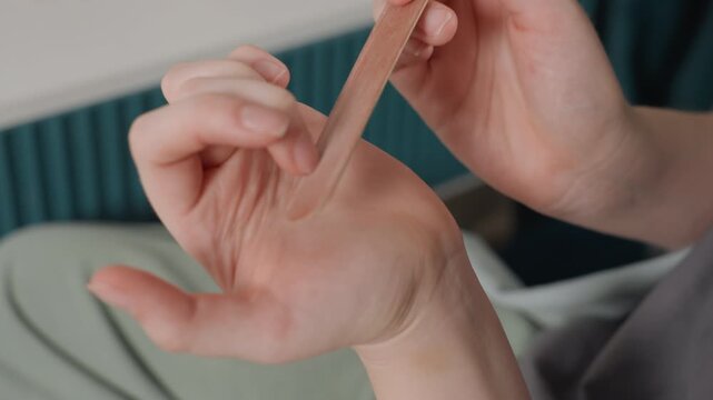 Closeup hand filing nails on bed, intimate athome manicure scene showing woman shaping thumbnail with emery board, gentle buffing of cuticles, natural light, soft bedding background, calm wellness