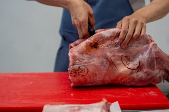 Gros plan sur un boucher en tablier bleu d&eacute;sossant avec pr&eacute;cision une &eacute;paule d'agneau sur une planche rouge, illustrant un savoir-faire artisanal et professionnel.