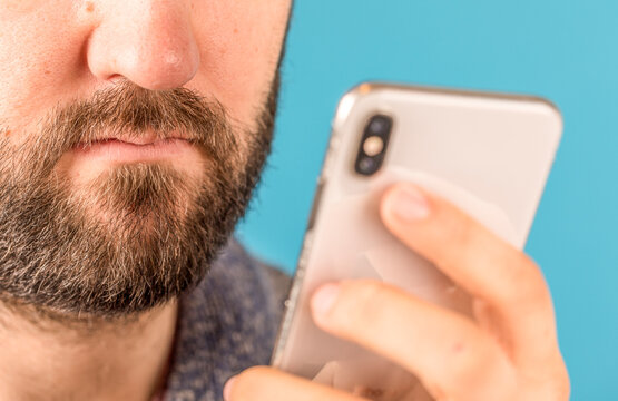 Close up shot of a bearded man holding a mobile phone in front of a blue background. Technology usag