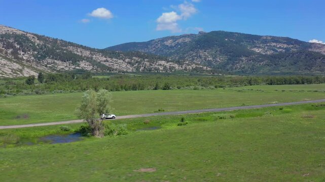 Aerial view of car driving slowly along quiet countryside road through green fields and mountains in Kazakhstan, peaceful travel concept, slow living lifestyle, calm nature escape and relaxing journey